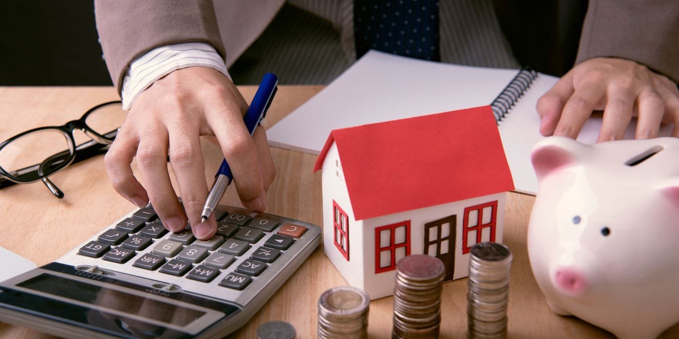 A person calculating housing costs, a small model house, and a piggy bank, symbolizing the real estate market differences between Edmonton vs Vancouver.