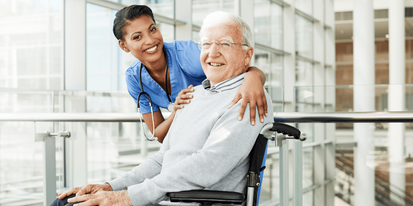 A smiling healthcare worker standing behind and embracing an elderly man in a wheelchair inside a modern medical facility, illustrating education and healthcare facilities when comparing living in Calgary vs Toronto.