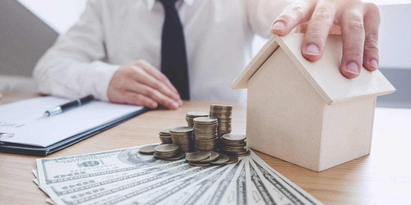 A person in a suit holding a small house model next to stacks of coins and dollar bills, symbolizing the cost of living in Montreal vs Toronto.