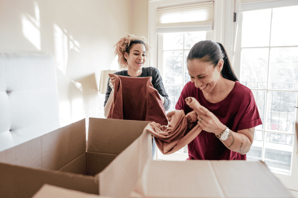 Two women happily unpacking a box in a bright room, excited about their move to Canada.