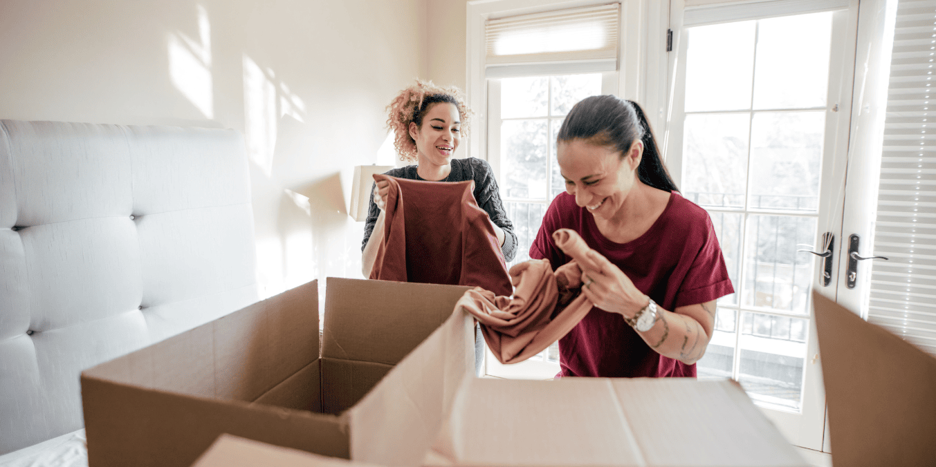 Two women happily unpacking a box in a bright room, excited about their move to Canada.