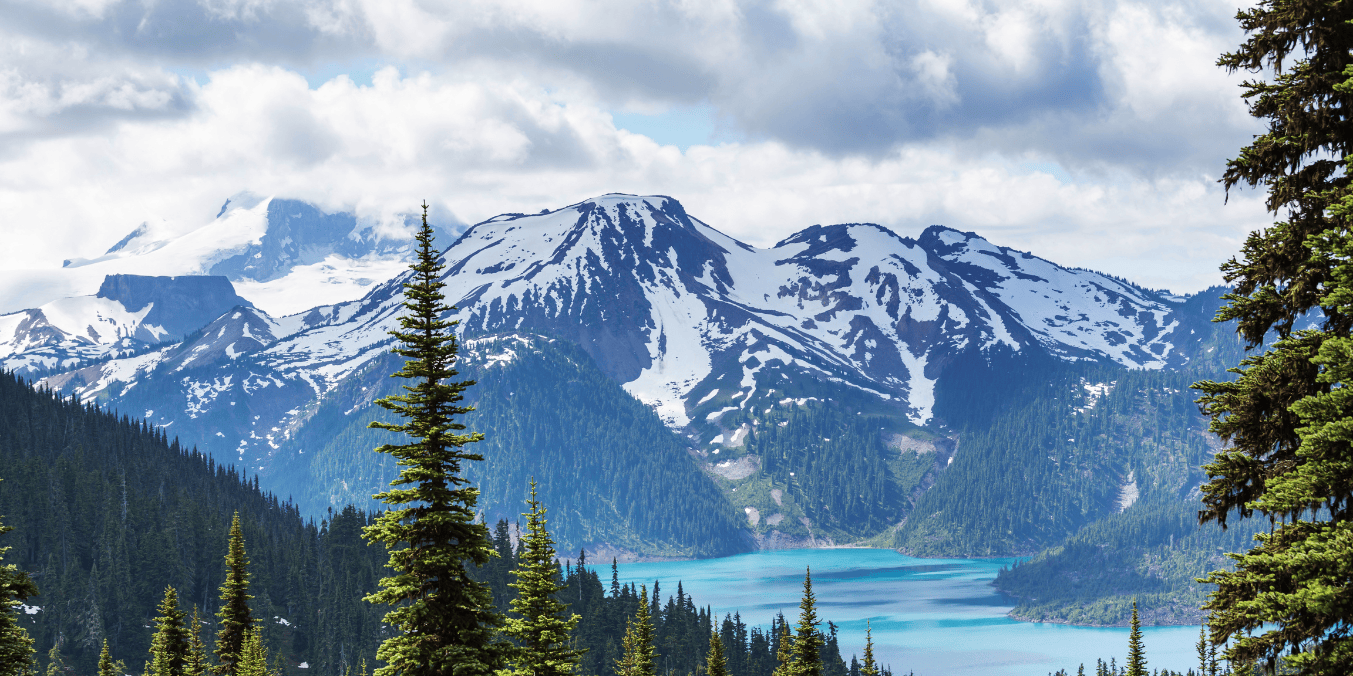 A breathtaking view of a snow-capped mountain range with a clear blue lake surrounded by lush pine trees, showcasing the stunning natural beauty that makes moving to Canada so appealing.