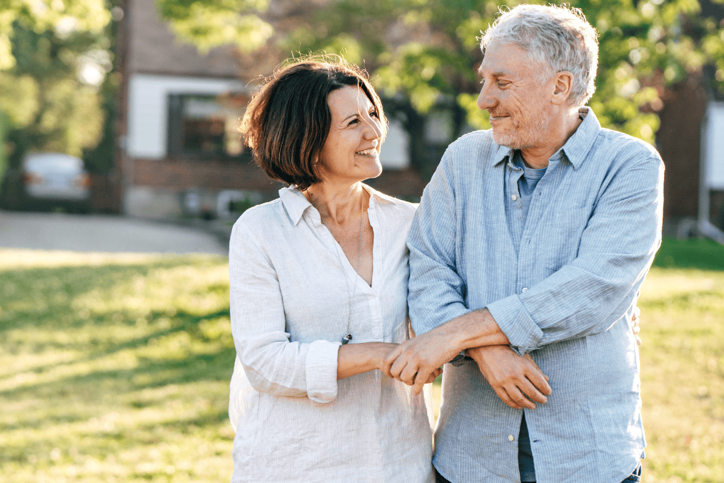 A smiling middle-aged couple stands arm in arm in a sunny suburban neighborhood, looking at each other affectionately, symbolizing the possibilities of starting a new life together in Canada. The image reflects the theme of the article, 