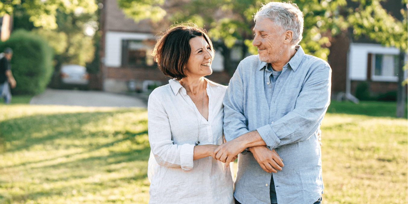 A smiling middle-aged couple stands arm in arm in a sunny suburban neighborhood, looking at each other affectionately, symbolizing the possibilities of starting a new life together in Canada. The image reflects the theme of the article, 