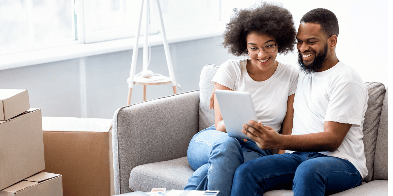 A couple smiling while researching how long spousal sponsorship takes in Canada on a tablet, surrounded by moving boxes.