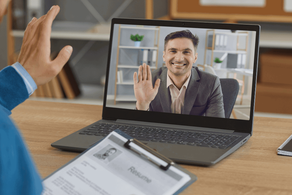 A man participating in an online interview, waving at a smiling interviewer on his laptop screen. The man holds a clipboard with a visible resume, symbolizing a virtual job interview at a Virtual Job Fair in Canada.