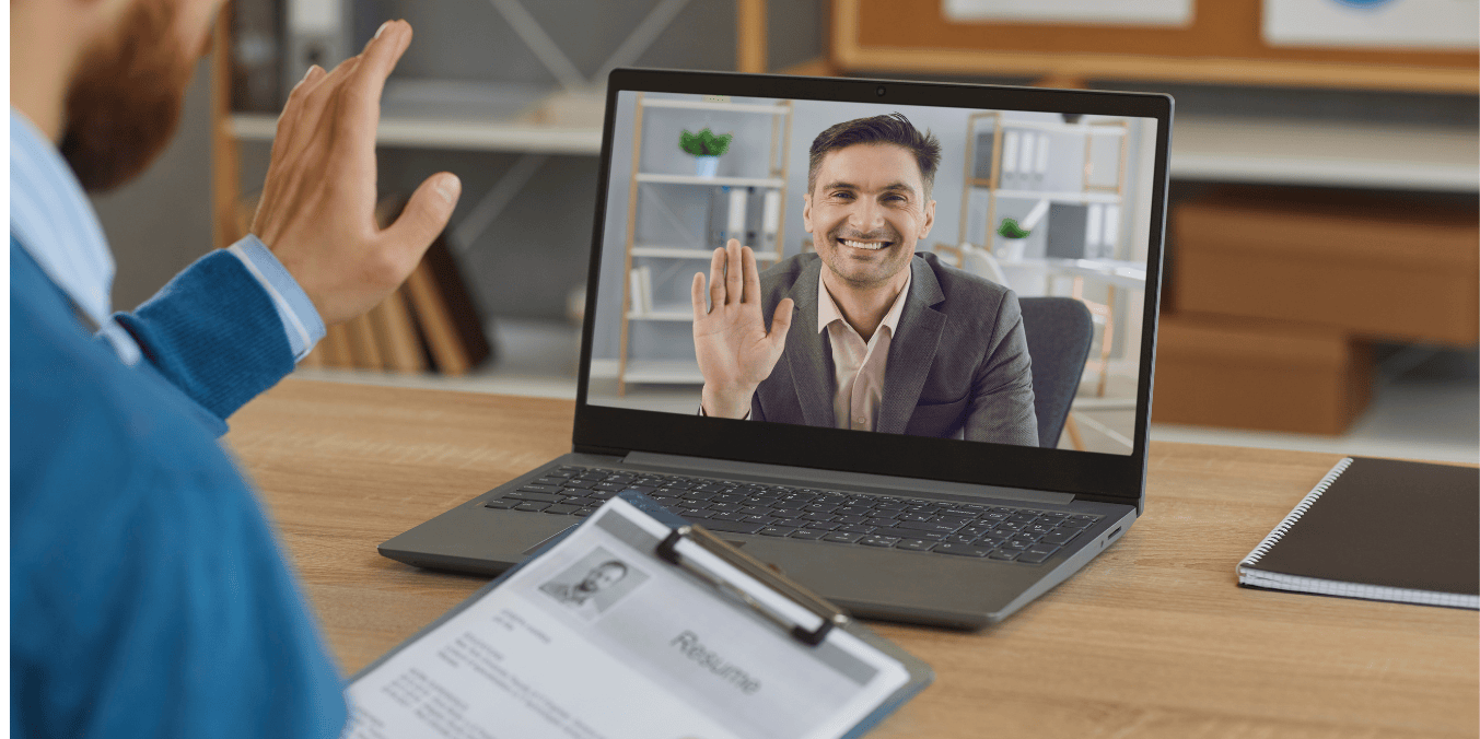 A man participating in an online interview, waving at a smiling interviewer on his laptop screen. The man holds a clipboard with a visible resume, symbolizing a virtual job interview at a Virtual Job Fair in Canada.