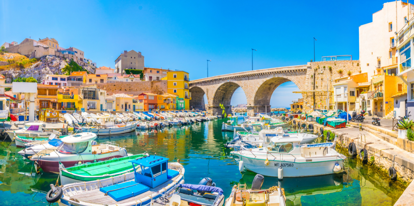 A picturesque marina scene in France, featuring colorful boats, pastel-colored buildings, and a historic stone bridge under a bright blue sky, showcasing the coastal charm and highlighting the lifestyle differences in life in Canada vs France.