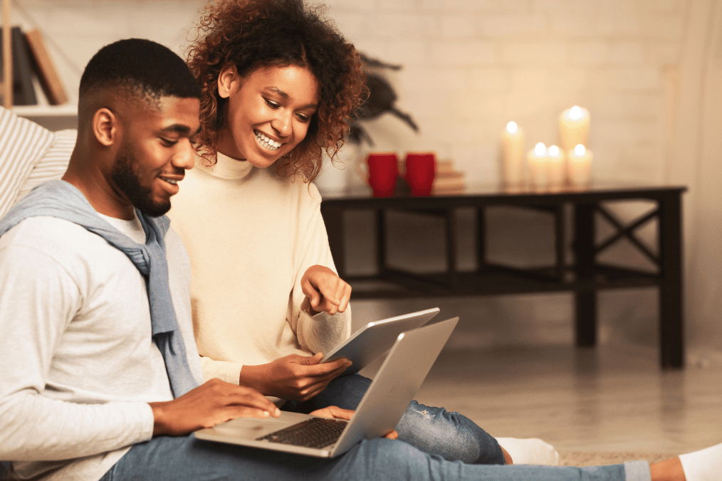 A happy couple sitting on the floor, using a laptop and tablet while discussing spousal sponsorship in Canada. Perfect for explaining, 