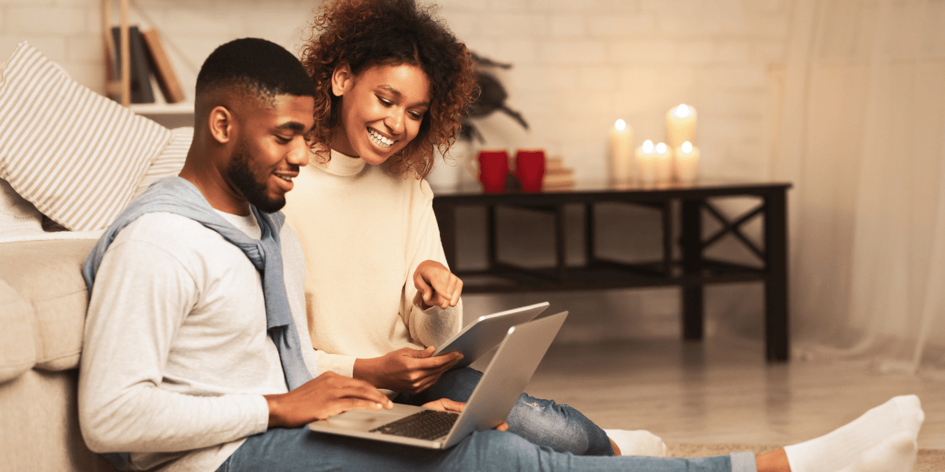 A happy couple sitting on the floor, using a laptop and tablet while discussing spousal sponsorship in Canada. Perfect for explaining, 