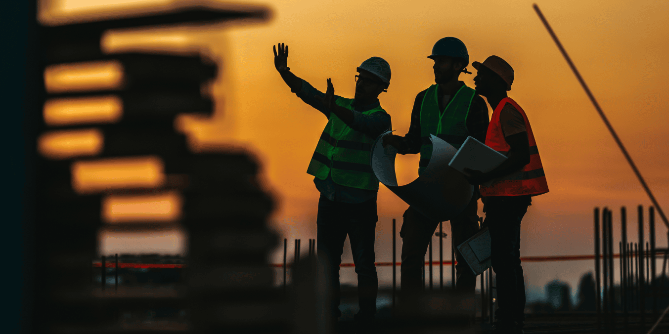 Three construction workers wearing safety vests and helmets review plans at a construction site during sunset, with one worker pointing ahead as they discuss the project.