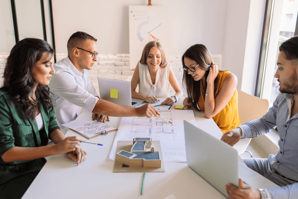 A diverse group of professionals discussing who can apply for BOWP while working on laptops and reviewing architectural plans in a modern office setting.