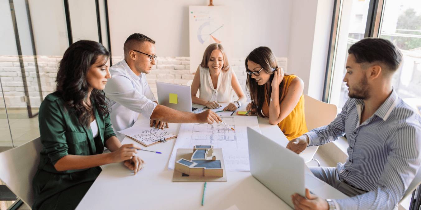 A diverse group of professionals discussing who can apply for BOWP while working on laptops and reviewing architectural plans in a modern office setting.